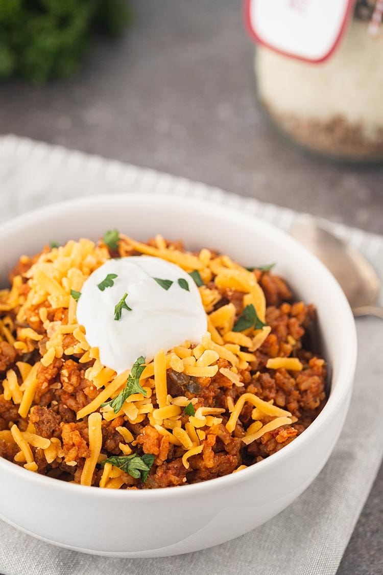 Shot of Beef Taco Rice Meal in a Jar made into a meal, in a white bowl, topped with shredded cheese and sour cream, garnished with fresh parsley.
