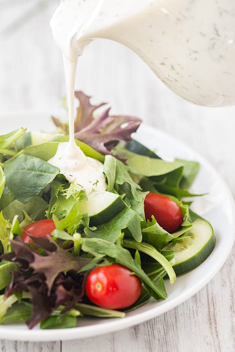 Picture of small glass pitcher of prepared ranch dressing being pour onto a basic green salad with tomatoes and cucumbers.
