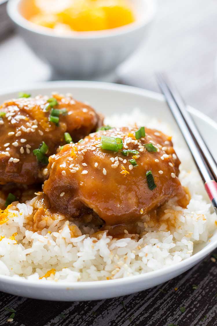 Sesame Orange Chicken thighs on a bed of rice, on plate with oranges in background.
