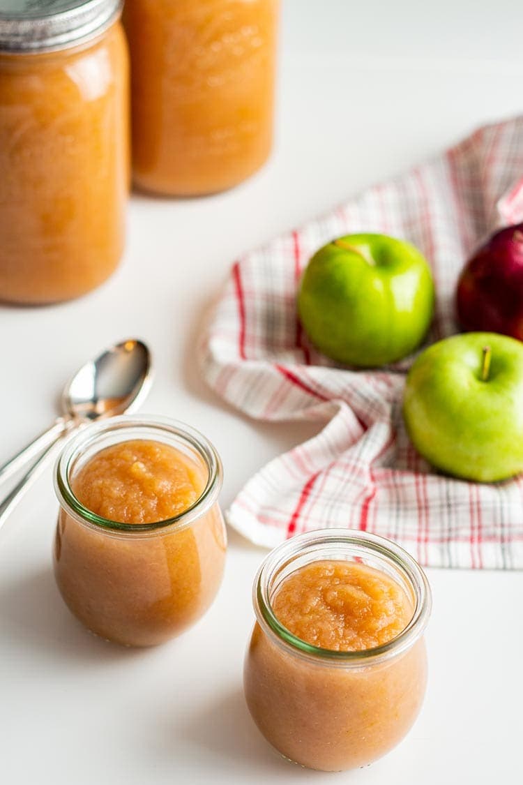 Jars of Instant Pot Apple Sauce on a white table with apples and towel in background.