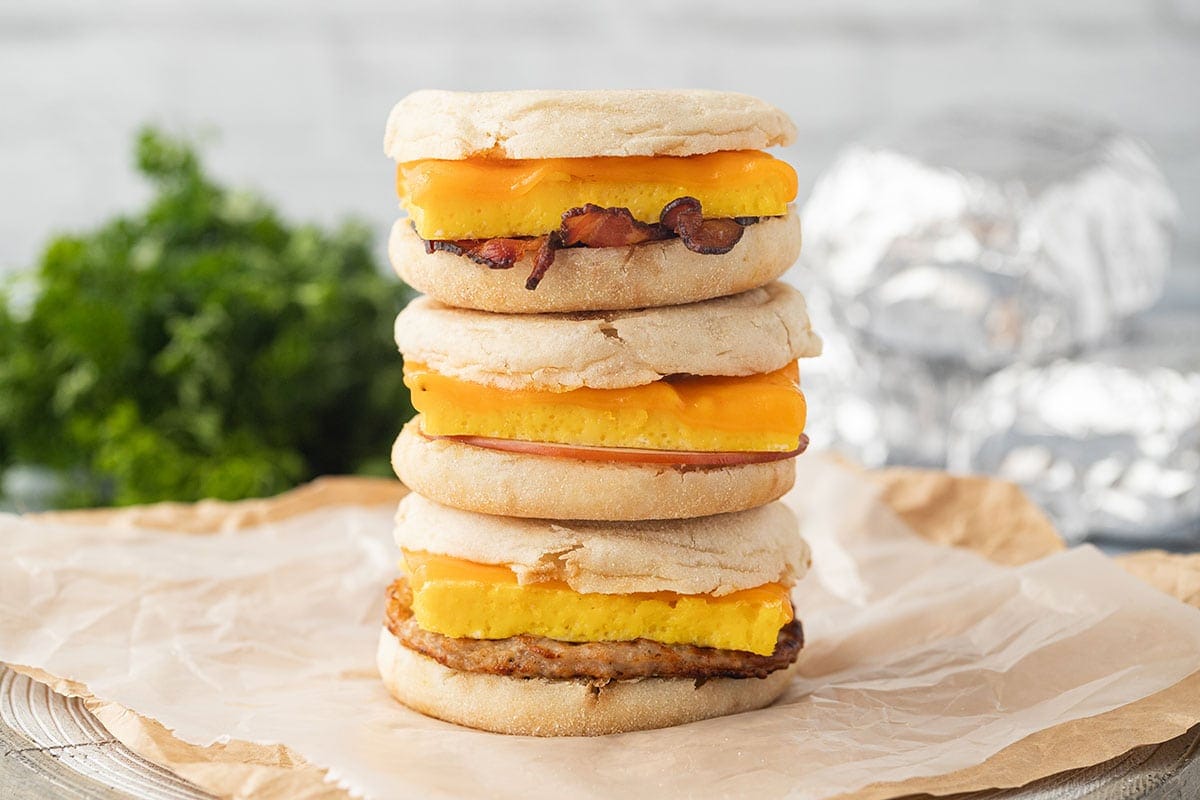A wide shot showing three stacked Freezer Breakfast Sandwiches with parsley and other wrapped sandwiches in the background.